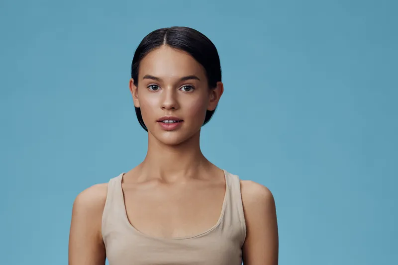 Young woman with a calm expression, wearing a beige tank top, posing against a light blue background Ideal for concepts like beauty and self confidence