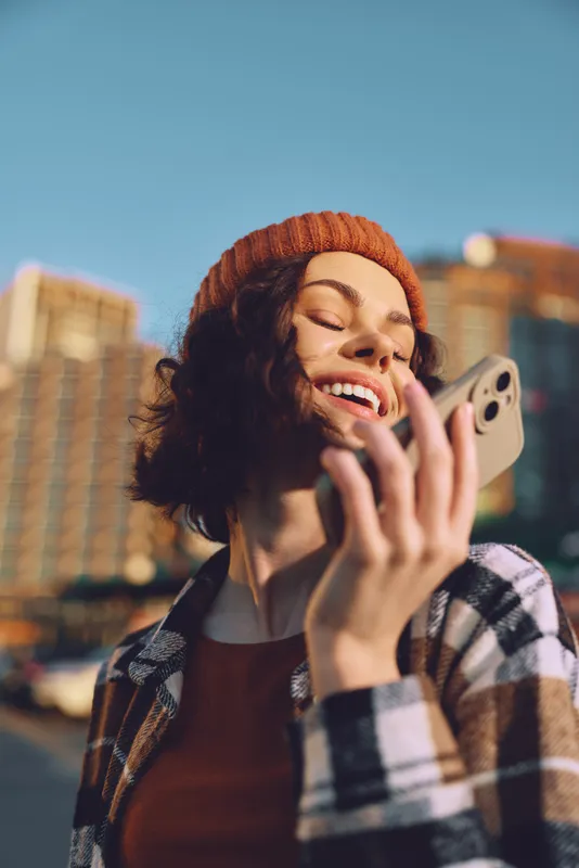 Woman with smile holding phone in city street wearing beanie, sunshine and candid urban portrait capturing authenticity, candid lifestyle and golden hour glow for mindful living and emotional
