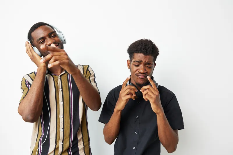 Two young African American men enjoying music, wearing headphones, expressing joy and energy. Their vibrant style reflects a modern urban lifestyle, highlighting fun moments together.
