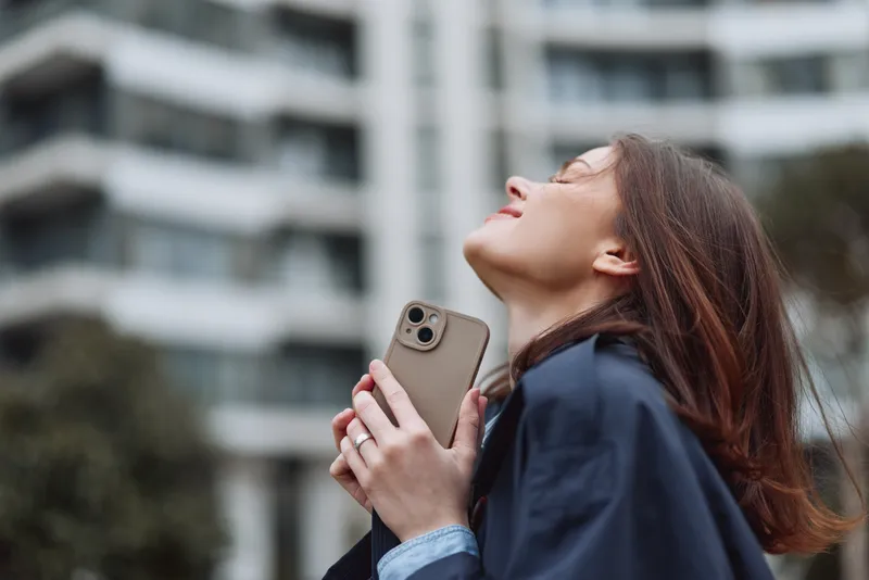Contemporary woman outdoors, looking up with phone in hand, sunlight on her face, urban backdrop, thoughtful mood, soft breeze, calm colors, open air and quiet atmosphere