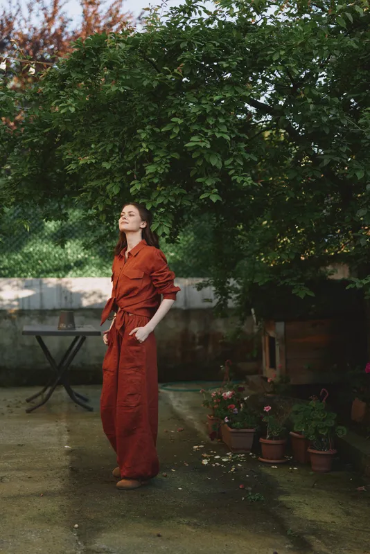 Woman in red jumpsuit stands in a garden amid trees and potted plants, looking upward with a calm, confident expression, capturing a candid lifestyle moment