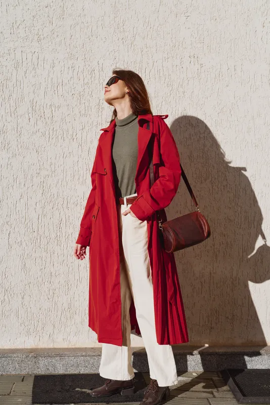 Young woman wearing a red trench coat and white pants stands confidently against a textured wall while looking upward with sunglasses on a sunny day.
