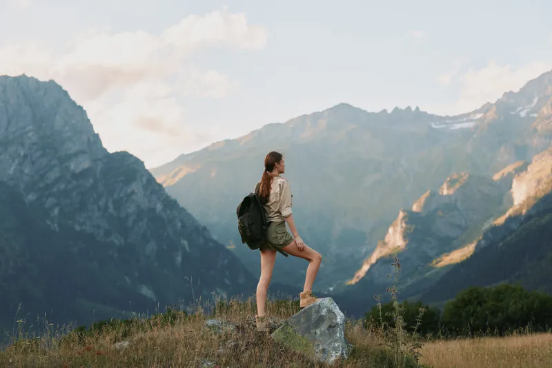 Young woman traveler with backpack stands on rock in mountain landscape enjoys nature and fresh air during summer hiking adventure outdoors.