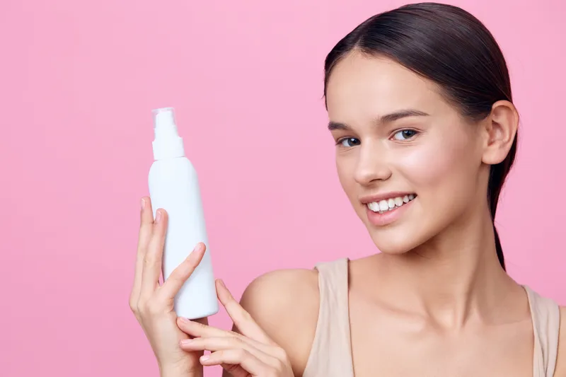 Young woman with a healthy glow holding a skincare bottle, smiling against a soft pink background, showcasing beauty and self care