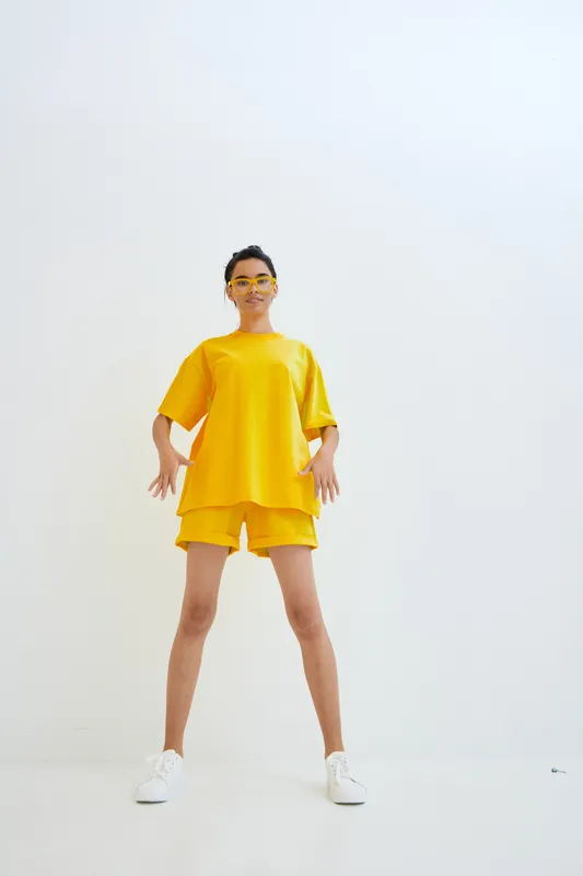 Young woman wearing bright yellow oversized t-shirt and shorts stands confidently against a white minimalist background, showcasing casual summer fashion and vibrant style.