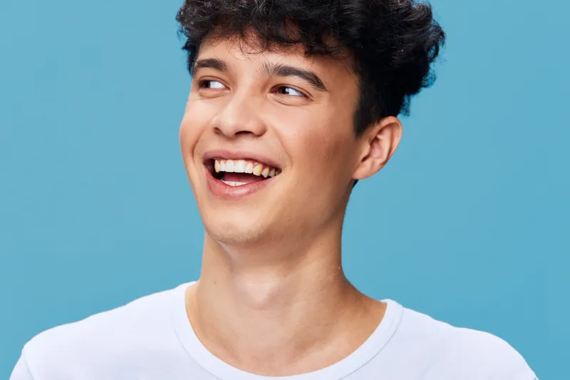 smiling man with curly hair wearing white t-shirt looking sideways on blue background, youthful expression, casual style, bright and cheerful atmosphere