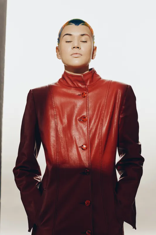 Young woman with closed eyes wearing a stylish red leather jacket, showing calm expression and modern short hair, standing against a plain white background.