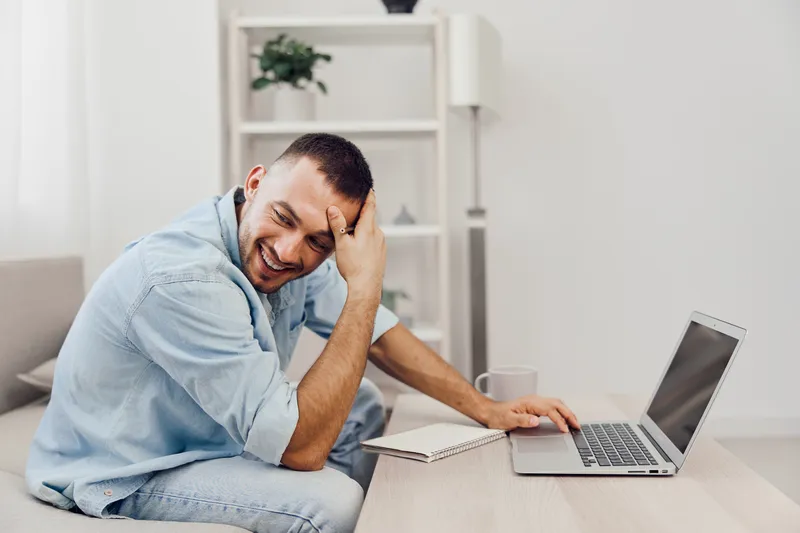 Smiling man working on laptop in a cozy, modern home office. Bright, welcoming atmosphere with soft colors and natural light. Perfect for remote work themes.