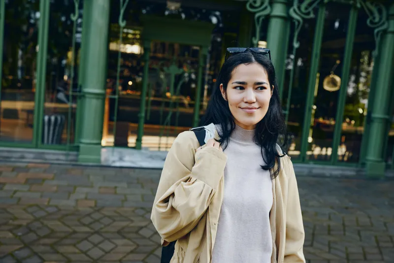 Woman with backpack wearing a beige coat and gray sweater outdoors on a city street, casual fashion style in an urban setting, candid moment with relaxed smile