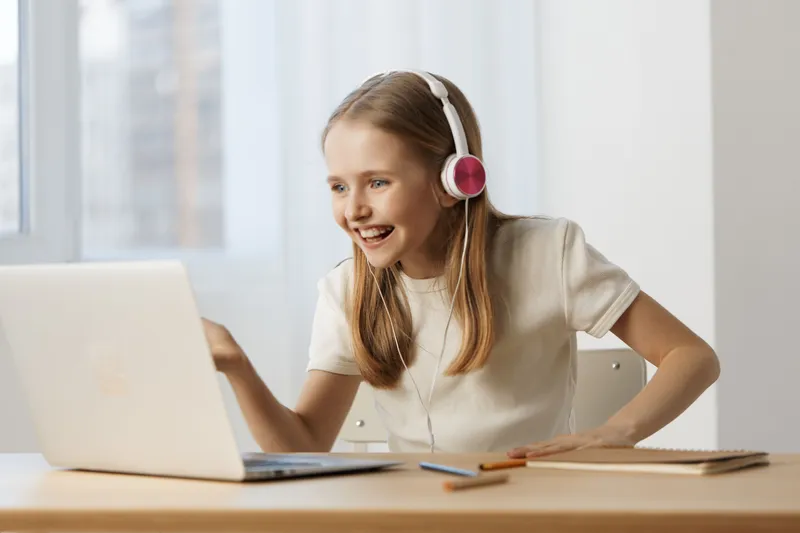 Happy girl enjoying online learning with headphones while sitting at a desk, wearing a casual white shirt, and smiling in a bright room with natural light