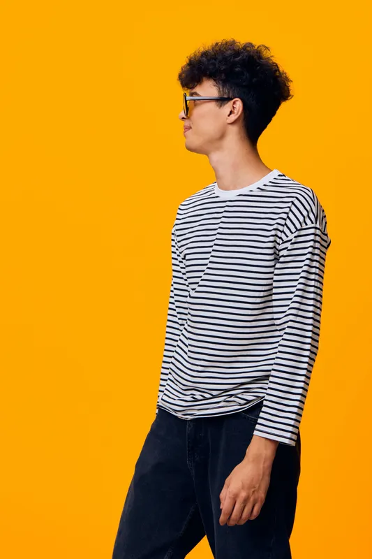 young man with curly hair wearing glasses and striped shirt stands sideways against vibrant orange backdrop, casual style and thoughtful expression in studio lighting