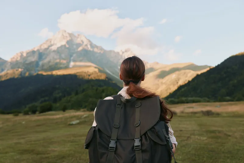 Woman traveler with backpack stands on green field looking at mountain peak under blue sky, enjoying outdoor adventure and nature lifestyle in scenic landscape.
