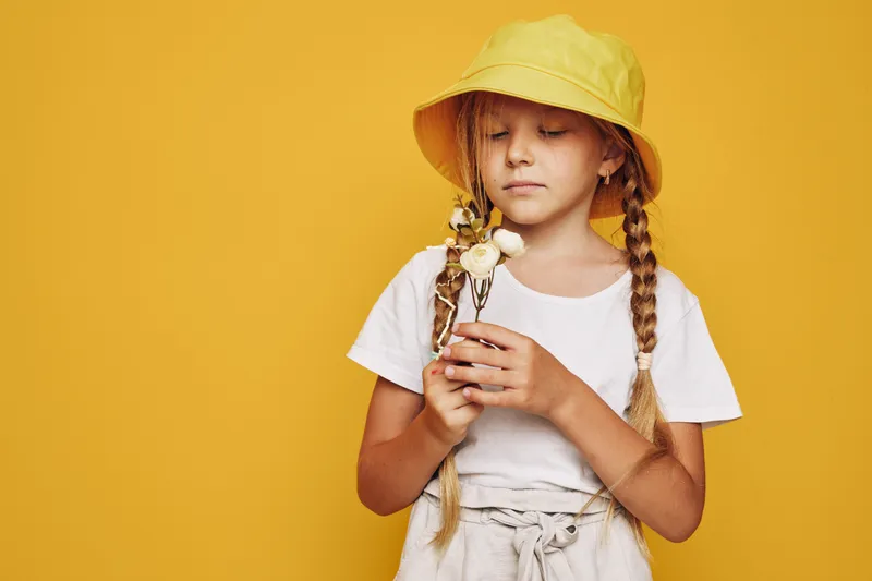 Young girl with braided hair in a yellow hat holds white flowers in both hands. She stands against a solid yellow background wearing a white shirt and beige pants. Concept of childhood and nature.