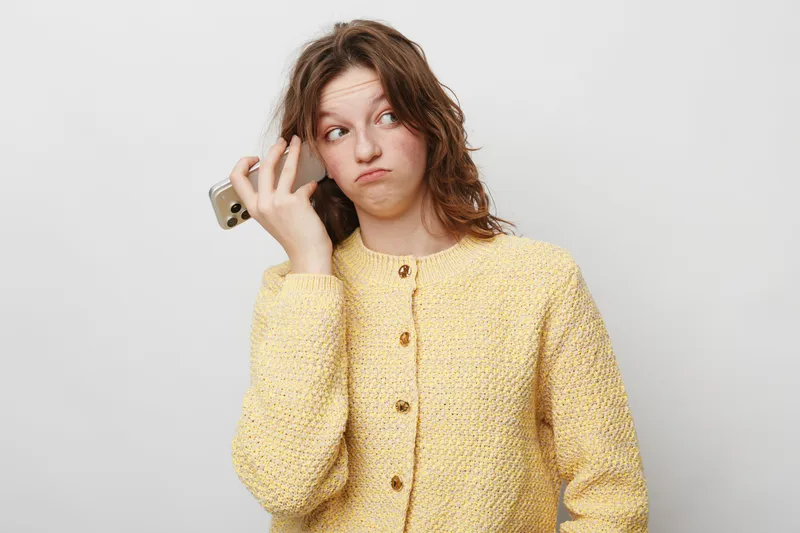 Young woman in yellow sweater holds smartphone near ear with doubtful expression, looking away. Isolated on white background. Concept of confusion, communication, and indecision.