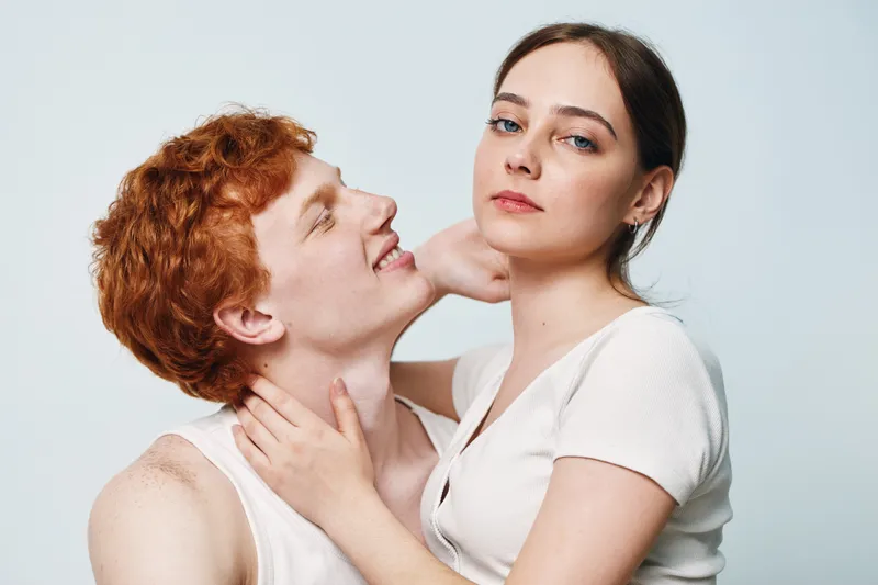 Couple, young man and woman with red hair and brown hair, intimate pose against white background. Expressions showing connection with skin and face closeup in studio light.