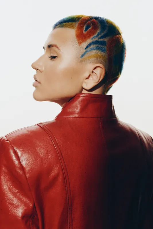 Woman with colorful artistic hairstyle and shaved side wearing a red leather jacket, showcasing modern edgy fashion and creative self-expression in a minimalist setting.