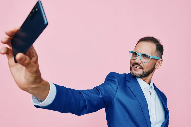 A professional man in a blue suit takes a confident selfie on a solid pink background, exuding energy and focus as he captures a moment of success.
