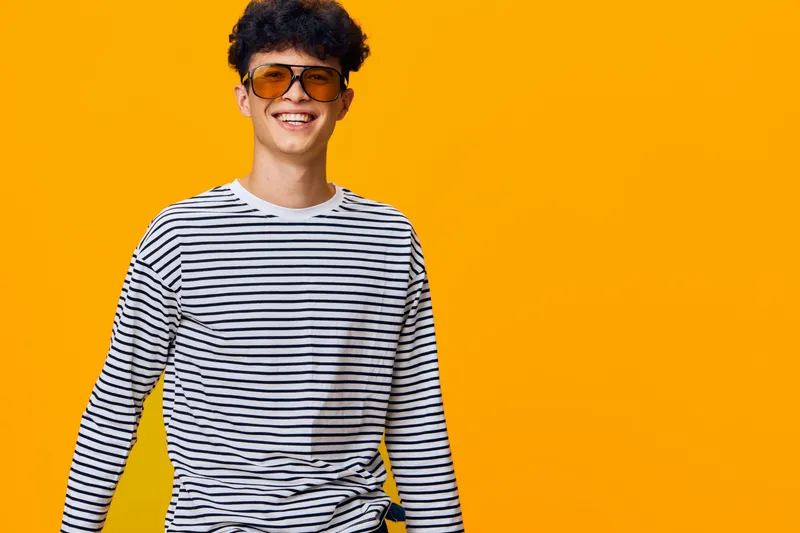 smiling man wearing striped shirt and sunglasses stands confidently against bright orange background, displaying joyful expression and casual style in vibrant setting.