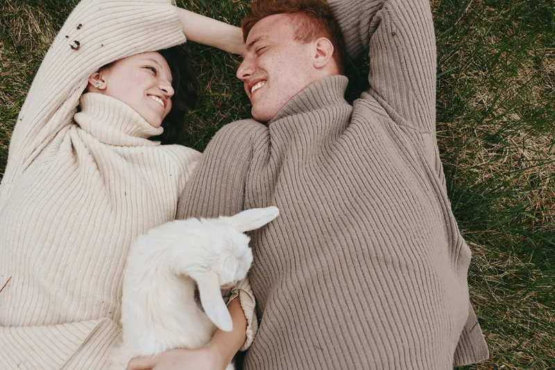 Smiling couple in cozy sweaters lying on grass with a white goat kid, enjoying a rustic outdoor moment. Lifestyle scene highlighting warmth, companionship, and nature, with soft daylight and gentle