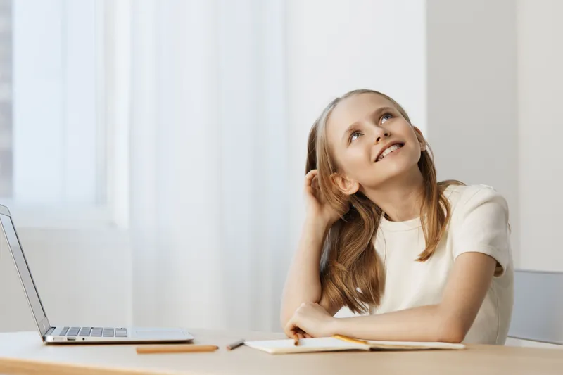 Thoughtful young girl sitting at a desk with a laptop, smiling and looking up, reflecting on ideas in a bright and minimalistic workspace