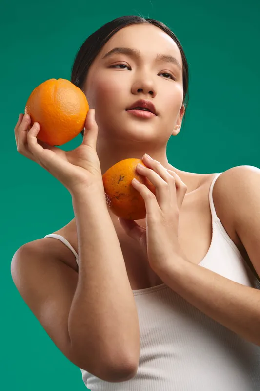 Asian woman holds bright oranges in a close up studio portrait against a teal backdrop, creating a cheerful mood and fresh energy, showcasing natural beauty and modern style.