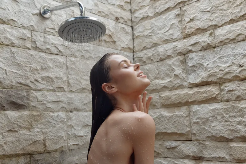 woman showering with closed eyes under rain showerhead, relaxing in natural stone tiled bathroom, wet hair and serene expression, calm mood