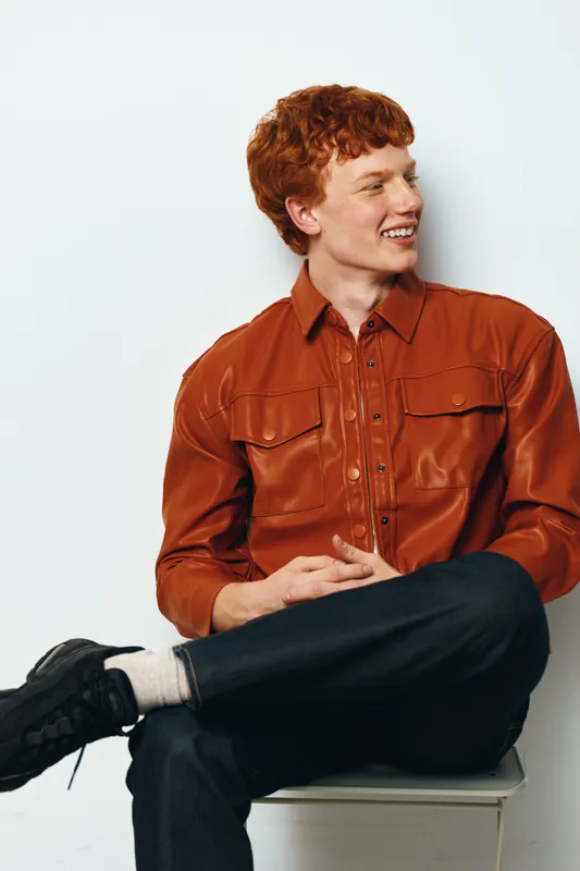 Young man with red hair wearing brown leather jacket and dark jeans sitting on chair smiling looking sideways isolated on white background in casual style portrait.