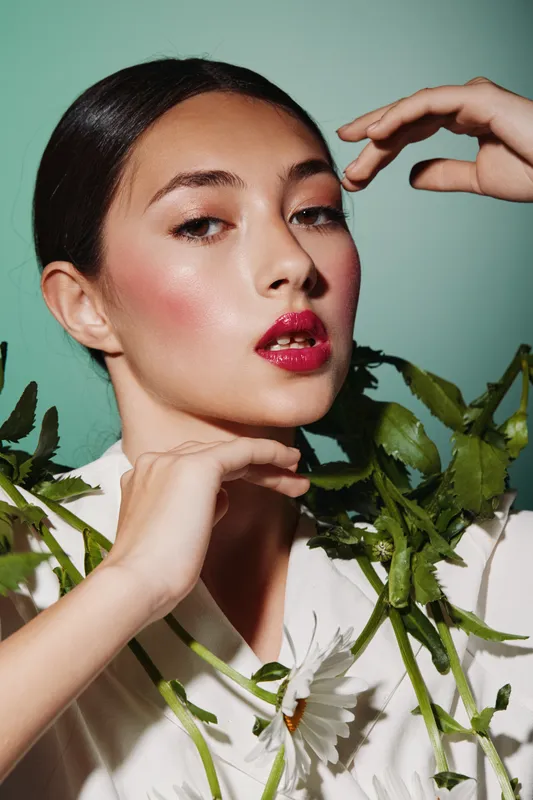 Young woman with glossy red lipstick and lush greenery poses with delicate hands and soft glancing eyes on pastel green background. Beauty fashion portrait concept.