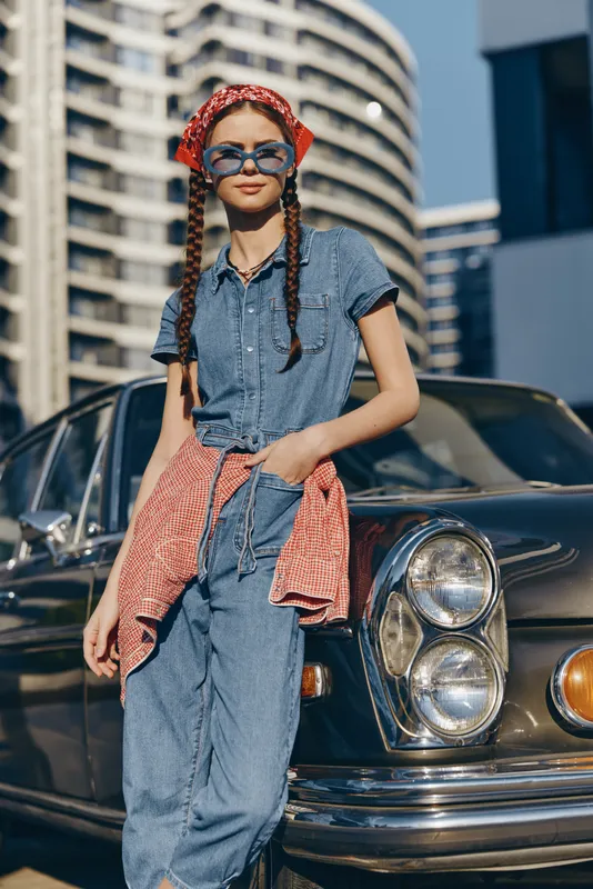 smiling woman in denim outfit with red bandana and sunglasses standing near classic vintage car in urban sunny setting on clear day