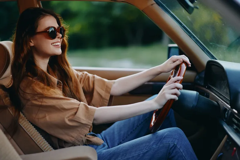 Happy young woman with long hair driving a vintage car on a sunny day, wearing sunglasses and a casual outfit, expressing joy and freedom.