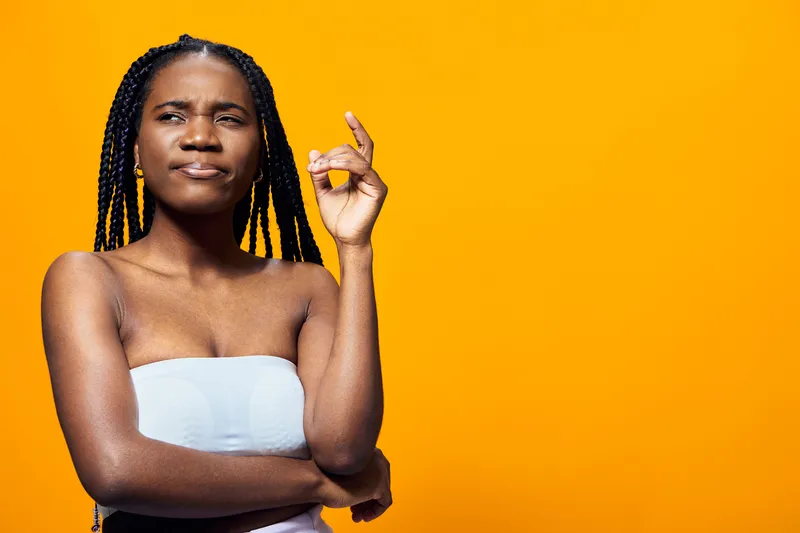 Thoughtful young Black woman with braids, wearing a light blue strapless top, pondering against a vibrant yellow background, expressing curiosity