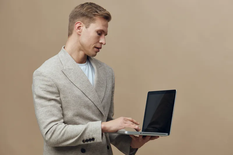 Professional young man with short blond hair, wearing a casual beige blazer, holding a laptop while standing against a plain beige background in a studio setting. Business, technology, workspace conce
