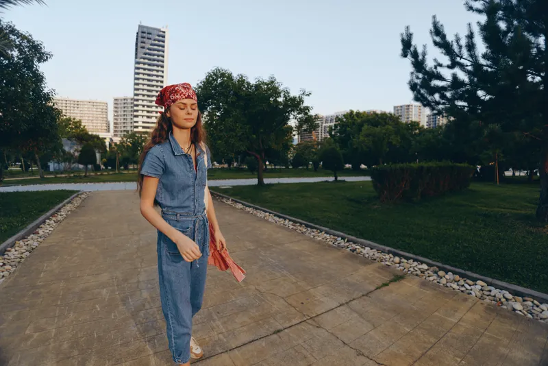 young woman walking outdoors wearing denim jumpsuit and red headscarf in urban park with green trees and modern buildings background