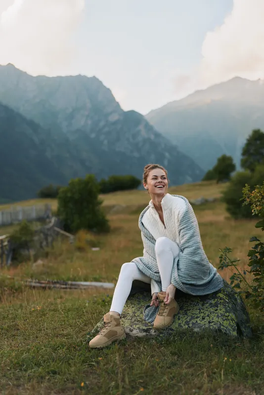 Smiling woman in cozy sweater sitting on rock in mountain meadow, autumn landscape, casual outfit, relaxed mood, natural light, outdoor lifestyle