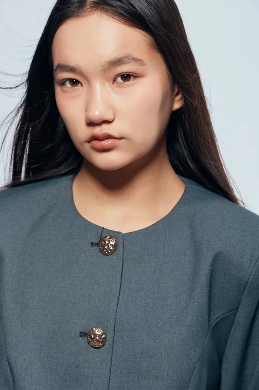 Young woman with long dark hair wearing buttoned gray jacket posing against light background. Serious expression, natural makeup, and casual hairstyle for portrait photography.