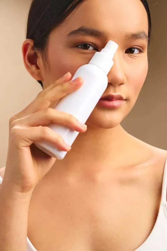 asian woman applies skincare product with a spray bottle in a minimal studio, her calm expression and soft lighting convey a clean, fresh beauty routine.