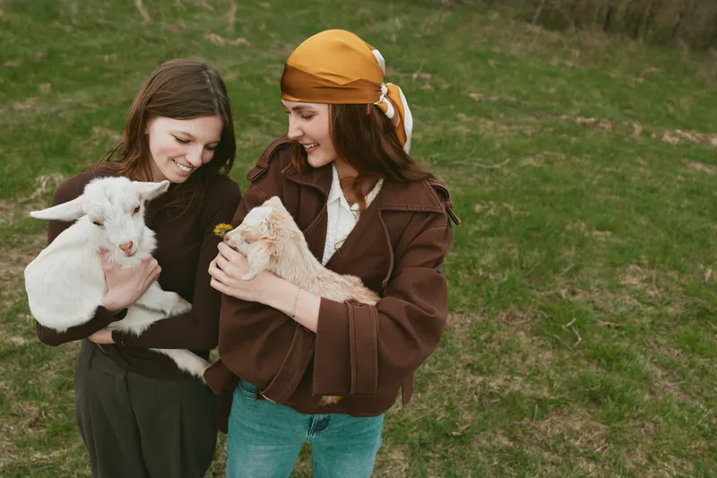 Two young women enjoy a lifestyle moment outdoors holding a white baby goat and a brown kid with smiles and casual autumn clothing on green grass in natural light.