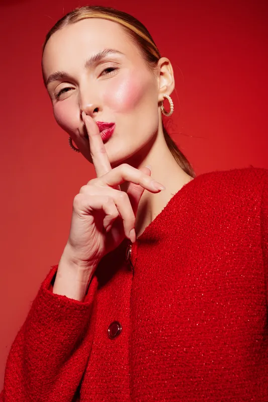 Young woman in red sweater with finger on lips makes shushing gesture on red background. Concept of silence, secrecy, and quiet communication with confident expression.