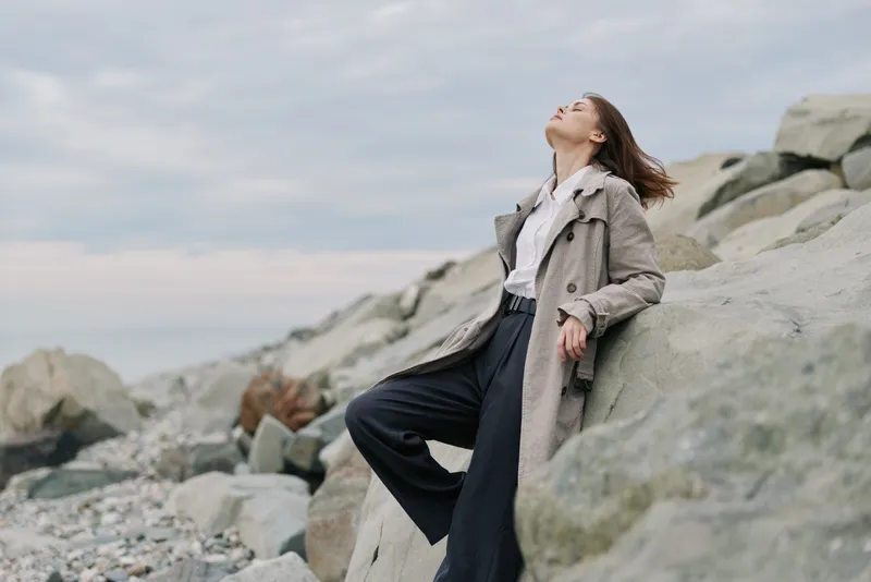 Young woman in a coat leans back on rocky coastline, eyes closed, enjoying fresh air and nature. Casual outfit and serene mood create a concept of relaxation and freedom outdoors.