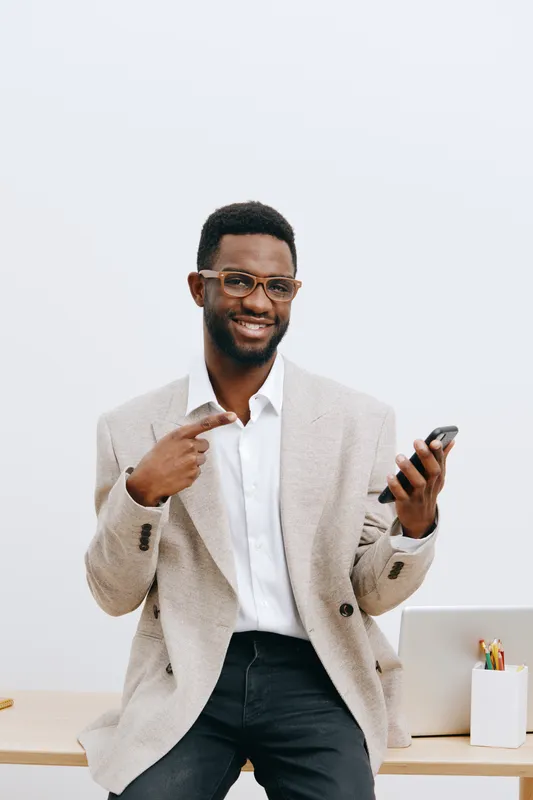 A full body portrait of a smiling cheerful young African American man wearing glasses, a beige blazer, and a white shirt, holding a mobile phone in his hand, standing against a plain white background 