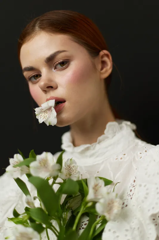 Young woman with long brown hair wearing a white lace dress, holding flowers, expressing tranquility and confidence, perfect for concepts of beauty and nature