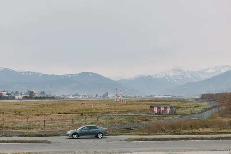 A gray sedan car parked on the roadside near an empty field with mountains in the background under a cloudy sky. Concept of travel, transportation, and nature landscape.
