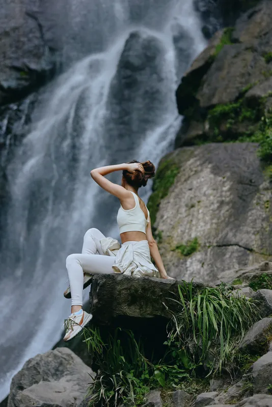 Young woman in white sportswear sits on rock near waterfall, enjoying nature, fitness, and outdoor adventure with lush greenery in the peaceful background.