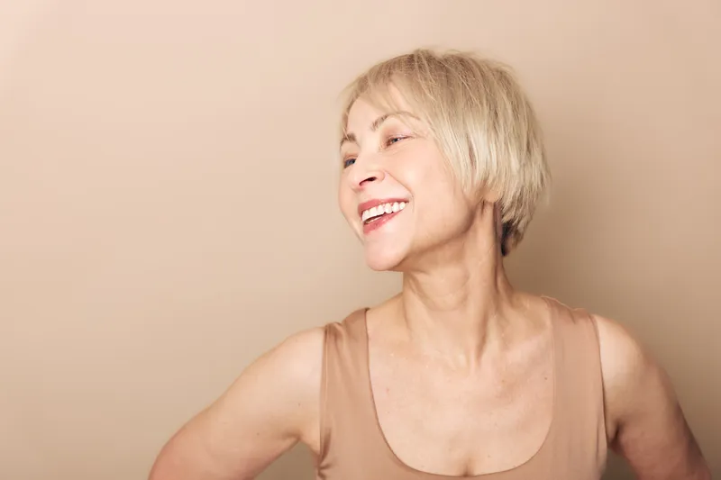 Smiling mature woman with short blonde hair wearing beige tank top looks to side against neutral background. Concept of happiness, aging, confidence, and natural beauty in studio portrait.