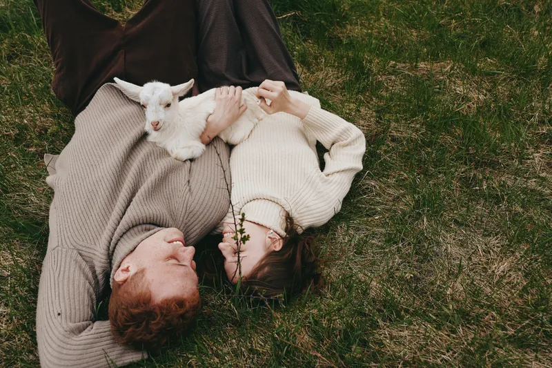 Couple lying on grass with a playful goat kid, cozy sweaters, and warm daylight. Outdoor lifestyle moment expressing affection, bond, and simple joy in nature. Warm and intimate scenery.