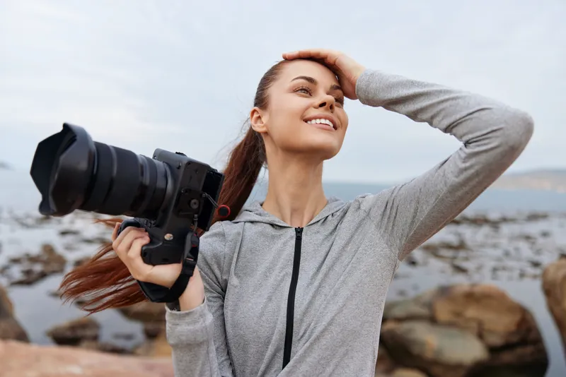 Happy young woman holding a camera on a rocky beach, dressed in casual sporty attire, capturing moments with a joyful expression, emphasizing creativity and outdoor adventure