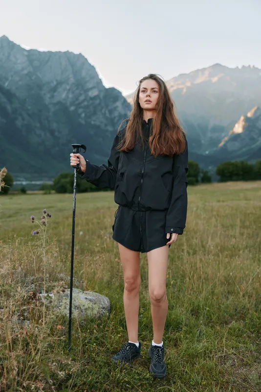 A focused young hiker stands in an open meadow with a trekking pole, framed by towering mountains. She looks confident and ready for adventure under a clear sky.