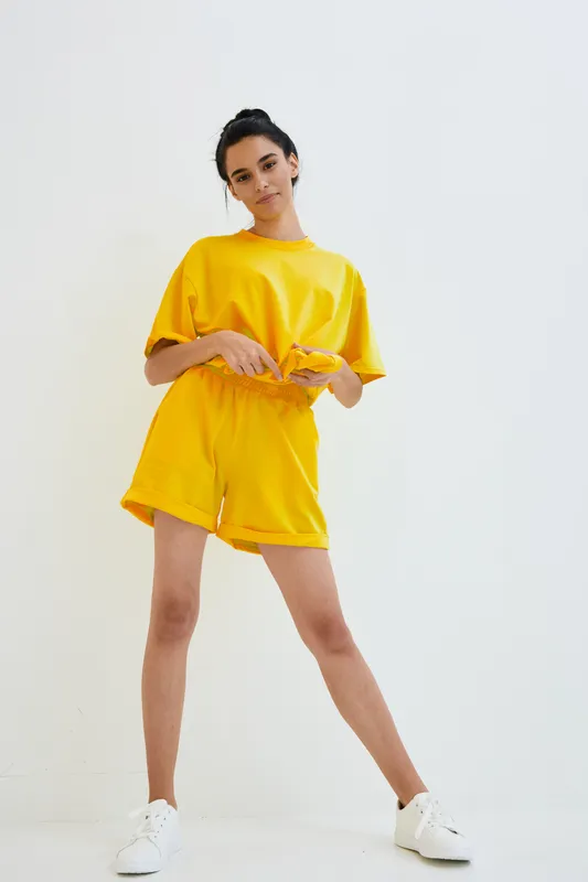 Young woman in bright yellow casual outfit stands confidently against white background, showcasing sporty and comfortable summer fashion with a relaxed mood.