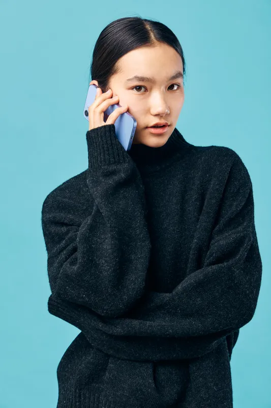 Young woman with black sweater holding smartphone to ear for communication and technology concept. Portrait with serious expression on blue background for conversation and connection.