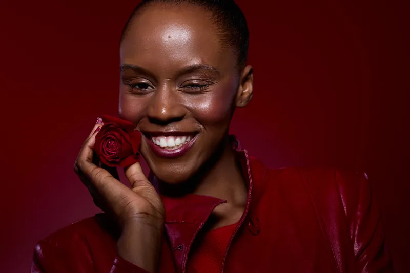 Smiling dark skinned African woman holds red rose near face wearing red outfit on red background. Concept of beauty, happiness, love, and floral aesthetics with natural skin glow.